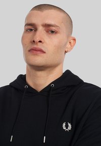 Sudadera negra con capucha y cordones, con puntas de metal plateadas, que presenta un logo bordado en blanco en el lado izquierdo del pecho. Textura suave.