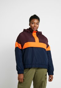Chaqueta con bloques de color que presenta una parte superior marrón, una franja media naranja y una sección inferior azul marino. El material parece suave, con capucha y puños elásticos.