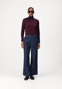 Maglione a collo alto a righe rosse e blu con maniche lunghe, abbinato a jeans scuri a gamba larga. Il modello indossa occhiali da sole neri e scarpe flat.