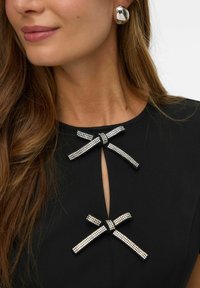 Czarna sukienka z dekoltem w kształcie dziurki, ozdobiona srebrnymi kokardkami pokrytymi kryształami. Gładki materiał, elegancki fason.