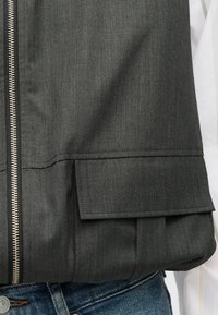 Šedá látková bunda so zapínaním na zips, textúrovaným povrchom a plochým vreckom na spodnej ľavej strane. Nosená cez rifľovinu.