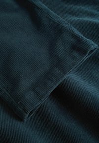 Tamno tirkizni kordurični materijal s izraženim vertikalnim rebrima i obrubljenim rubom, koji prikazuje mekanu teksturu i dosljedno uzorkovanje kroz cijeli materijal.