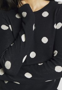 Mustat neulepusero, jossa on valkoisia pilkkuja. Valmistettu neuloksesta. Ominaisuuksiin kuuluu pitkät hihat ja pyöreä pääntie, joka korostaa pilkkukuviota.