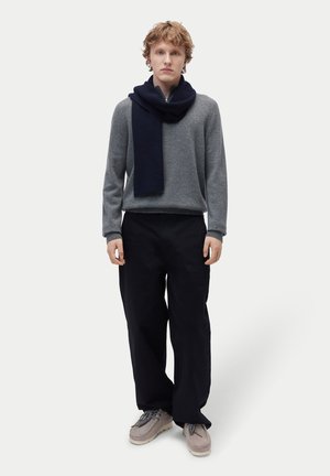 Junge Person steht und trägt einen grauen Pullover, marineblauen Schal, lockere schwarze Hose und beigefarbene Schuhe vor weißem Hintergrund.