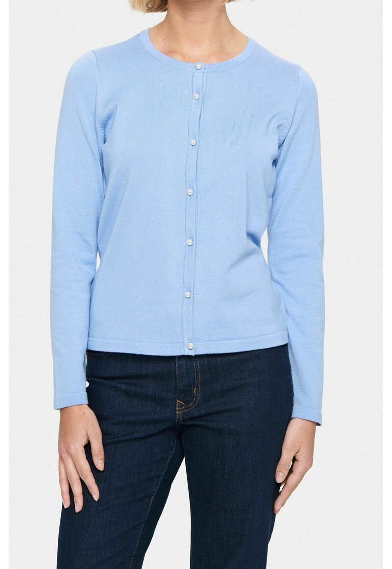 Femme portant un cardigan bleu clair boutonné avec des boutons en perle et un jean bleu foncé, montrée du cou aux hanches.
