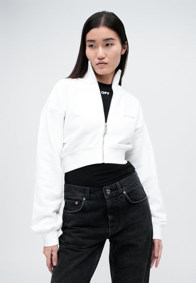 Felpa bianca cropped con zip realizzata in tessuto morbido, caratterizzata da maniche lunghe e colletto alto. Indossata sopra una maglietta nera e jeans scuri.