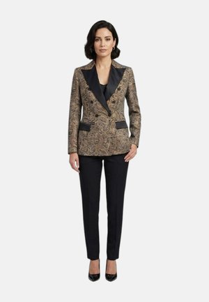 Donna che indossa un blazer con motivo paisley con rever neri, pantaloni neri e tacchi neri a punta, in piedi davanti a uno sfondo semplice.