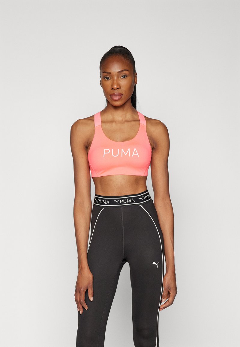 Rožinė sportinė liemenėlė su plačiomis petnešomis ir balta "PUMA" logo. Suderinta su juodomis aukšto liemens legging'omis, turinčiomis baltus apvadus ir logotipą.