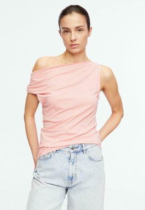 Donna che indossa un top monospalla rosa chiaro e jeans azzurri, in piedi con le mani in tasca davanti a uno sfondo bianco semplice.