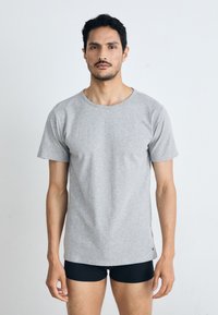 Férfi szürke póló, rövid ujjú, kerek nyakkal, puha anyagból, karcsú fazonban. Fekete rövidnadrág látható alul, minimalista dizájnnal, minták nélkül.