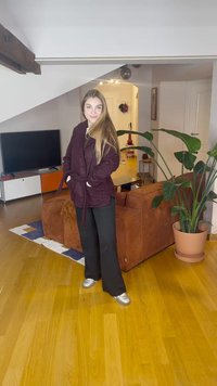 Burgunderfarget teksturert kåpe med belte, kombinert med svarte bukser og sølvfargede joggesko. Innendørs setting med en potteplante og sofa.