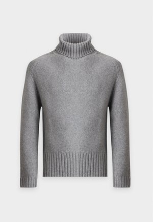 Homme portant un pull à col roulé gris, un jean bleu large et des chaussures noires, debout sur un fond blanc uni.