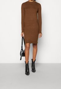 Abito lungo in maglione marrone con scollatura a coste, forma aderente, spacchi laterali, abbinato a stivaletti neri alla caviglia e una borsa piccola.
