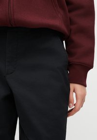 Άτομο με μαύρο παντελόνι και μπορντό μακρυμάνικη μπλούζα με ριμπ μανσέτες, το χέρι χαλαρό στο πλάι μπροστά σε απλό φόντο.