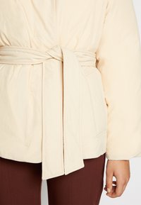 Casaco acolchoado de cor creme, com cintura cingida por um cinto, costura visível e bolsos laterais, combinado com calças castanhas escuras.