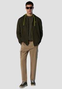 Felpa con cappuccio verde scuro con dettagli verde lime, abbinata a pantaloni cargo beige con tasche laterali e sneakers nere.