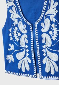Gilet en tissu bleu avec broderie florale et feuillage blanche symétrique le long des bords et des panneaux avant.