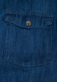 Tessuto in denim blu scuro con una tasca a rovescio dotata di chiusura a scatto in metallo, texture visibile e dettagli di cucitura lungo i bordi.
