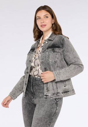 Mujer vestida con una chaqueta de denim gris con tachuelas y pantalones de cintura alta a juego, con una blusa estampada debajo, de pie frente a un fondo claro.