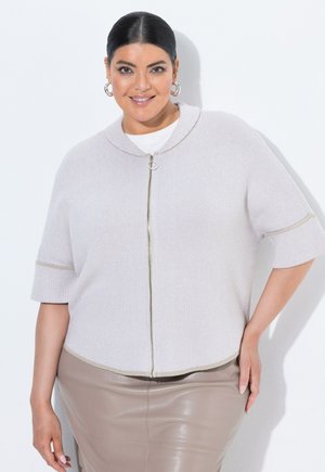 Lachende vrouw met donker haar draagt een lichtgrijze korte mouw ritsvest en een beige leren rok, poserend tegen een witte achtergrond.