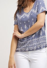 Krátký rukáv top v měkké, texturované modré tkanině s bílými geometrickými vzory, kulatým výstřihem a volným střihem.