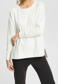 Sudadera blanca de manga larga con un corte relajado, cuello redondo, aberturas en los laterales y puños acanalados, combinada con pantalones negros. Textura de tela suave.