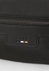 Borsa in tessuto nero con una patta in pelle liscia che presenta il logo "BOSS" e tre accenti colorati: nero, bianco e arancione. Chiusura con zip.