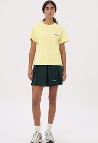Geel katoenen t-shirt met korte mouwen, gecombineerd met een donkergroene geplooide rok met witte branding. Casual schoenen maken de look compleet.