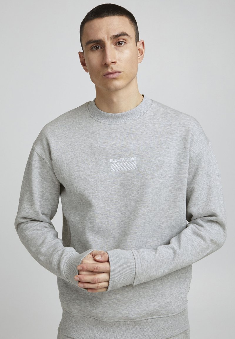 Sweatshirt gris en tissu doux, avec un col rond, des manches longues et un texte brodé blanc "SLD-EST.1999" sur le devant.