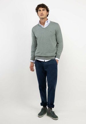 Hombre de pie con un jersey de cuello en V de color gris claro sobre una camisa blanca, pantalones azul oscuro, zapatos grises y calcetines con estampado sobre un fondo liso.