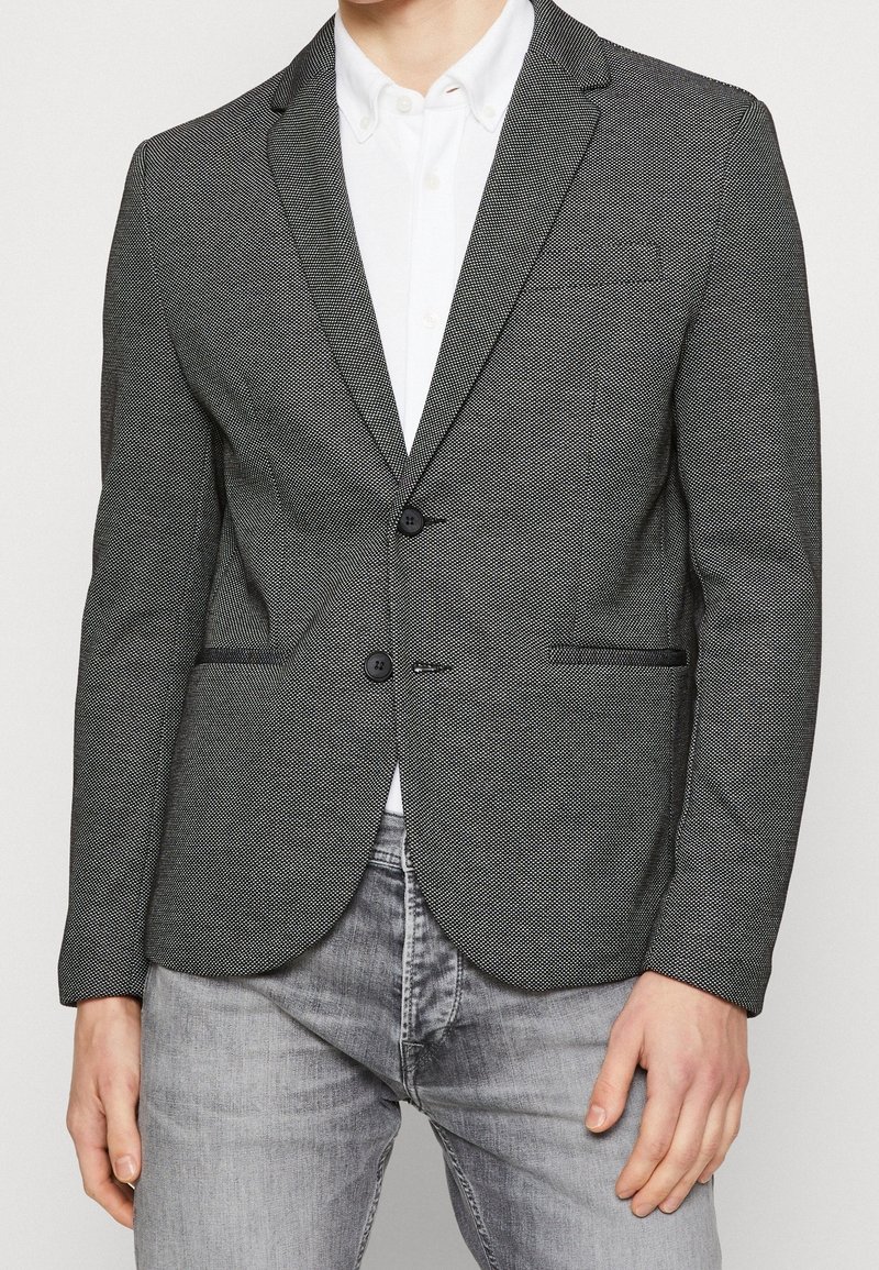 Czarna i biała marynarka w wzory z zaokrąglonymi klapami, dwoma przednimi kieszeniami i dwoma guzikami. Zestawiona z białą koszulą i szarymi dżinsami.