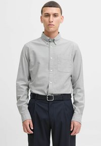 Camicia button-down in tessuto grigio chiaro, dotata di taschino sul petto, maniche lunghe e colletto. Abbinata a pantaloni blu scuro e una cintura nera.