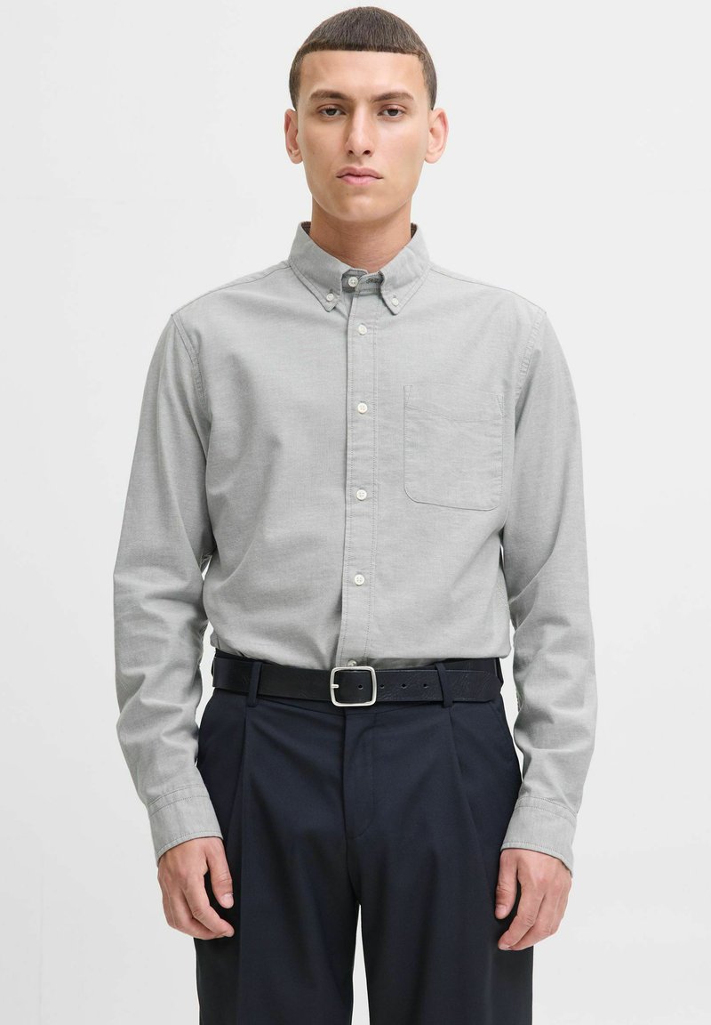 Camicia button-down in tessuto grigio chiaro, dotata di taschino sul petto, maniche lunghe e colletto. Abbinata a pantaloni blu scuro e una cintura nera.