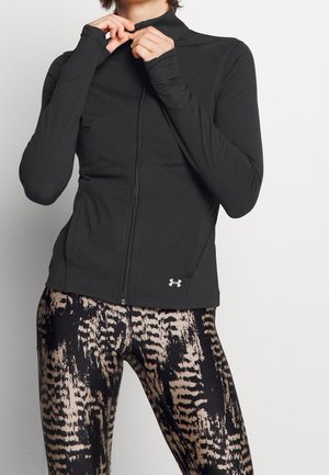 Person mit schwarzer Sportjacke mit Reißverschluss und gemusterten Leggings, Hände am Kragen, steht vor einem schlichten, hellen Hintergrund.