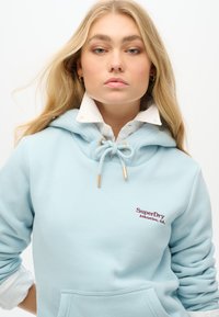 Sudadera azul claro con capucha ajustable, detalles en dorado y un bolsillo frontal. Presenta un logotipo bordado de "SuperDry Athletics, CA".