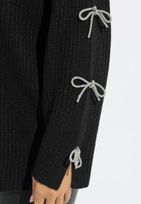 Czarny sweter dziany z srebrnymi, ozdobnymi kokardami na rękawach, prążkowaną strukturą i bocznymi rozcięciami.