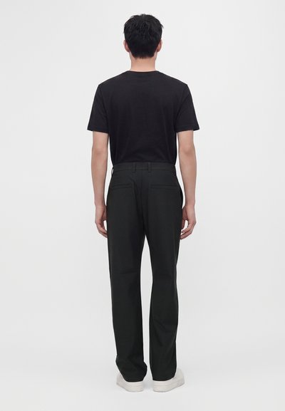 Homme debout, de dos, portant un t-shirt noir, un pantalon noir avec des poches arrière et des baskets blanches sur un fond blanc uni.