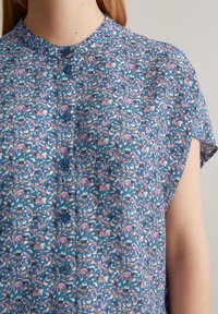 Button-up blouse in blauwe stof, met een bloemmotief met roze, groene en witte accenten. Korte mouwen en een bandkraag.