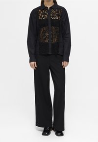 Chemise noire boutonnée avec panneau avant en dentelle, manches longues et col structuré, associée à un pantalon noir à jambes larges et des chaussures noires brillantes.