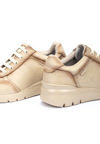 Zapatillas de cuero beige con cordones blancos, cuello acolchado, lados texturizados y suela redondeada. Presentan una marca discreta en el lateral.