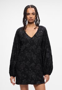 Melnas ziedu raksta kleitas ar V veida kakla izgriezumu, garām pūtīšu piedurknēm un teksturētu audumu. Ievērojama brīva piegriezuma un ceļgala garuma dizains.