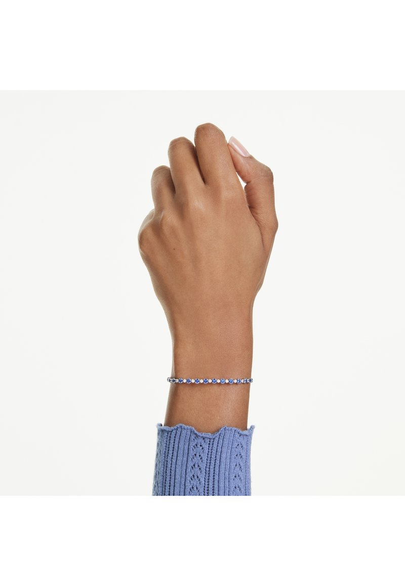 Bracciale d'argento con pietre blu, caratterizzato da un delicato design a catena. Indossato su una mano, contrasta con un maglione di lana blu a trama.