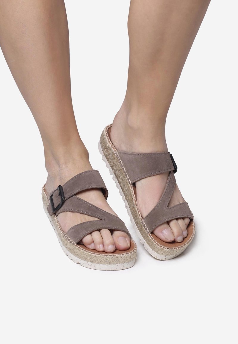 Pieds portant des sandales en daim taupe avec des lanières croisées, une boucle noire et des semelles plateforme épaisses tissées sur fond blanc.