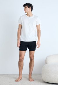 White cotton t-shirt and black shorts, fitted design. Model stands on a light carpet, barefoot, with a neutral background.