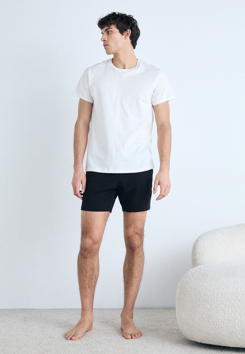 White cotton t-shirt and black shorts, fitted design. Model stands on a light carpet, barefoot, with a neutral background.