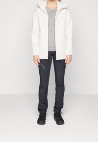 Personne portant une veste à capuche blanche, un t-shirt gris, un pantalon slim noir avec logo, et des chaussures de randonnée noires, debout devant un fond uni.