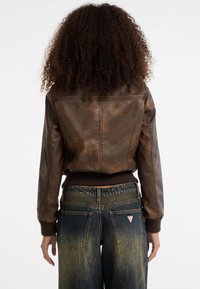Bruine leren bomberjack met een geribbelde zoom, weergegeven vanuit de achterkant. Gestyled met vervaagde denim jeans met pocketdetail.