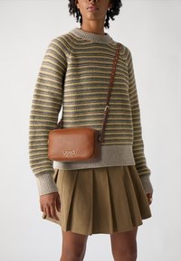 Borsa a tracolla in pelle marrone con logo dorato, indossata sopra un maglione a righe grigie e gialle, abbinata a una gonna verde oliva plissettata.
