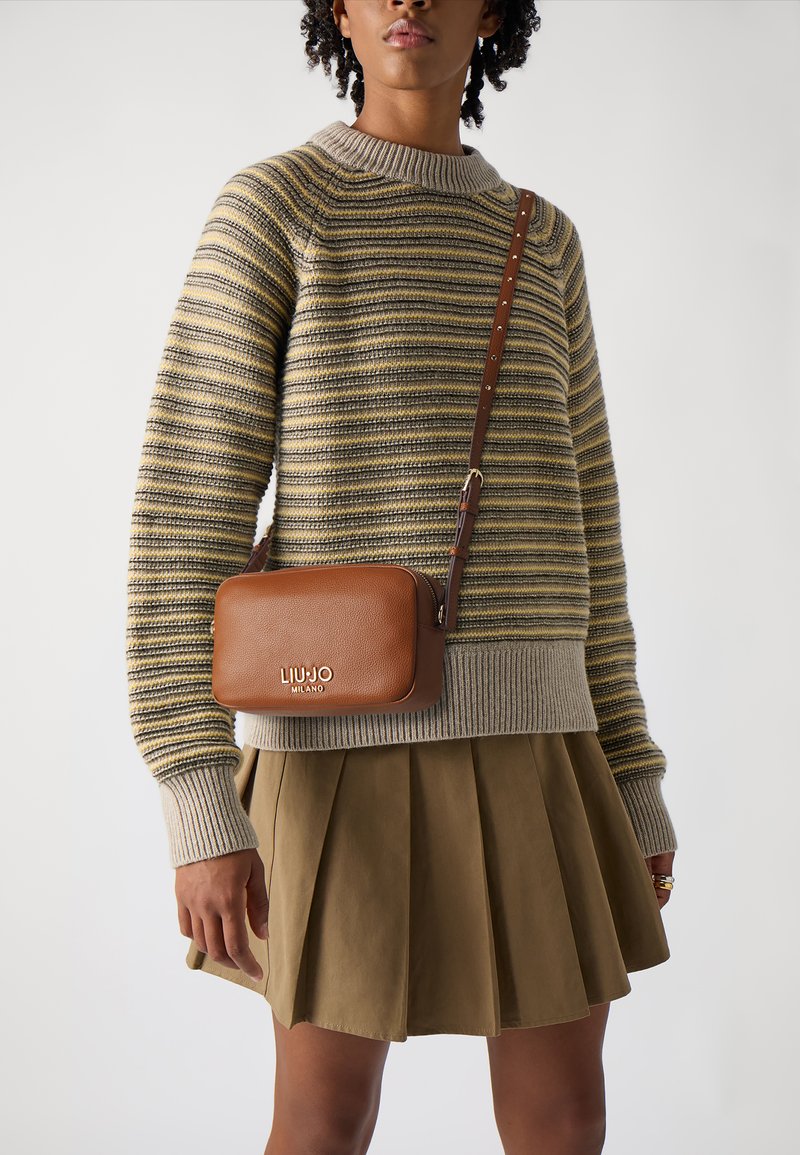 Borsa a tracolla in pelle marrone con logo dorato, indossata sopra un maglione a righe grigie e gialle, abbinata a una gonna verde oliva plissettata.