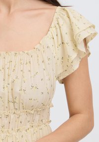Femme portant un chemisier jaune clair à manches courtes avec un petit motif floral, un col smocké avec élastique et des détails froncés à la taille.